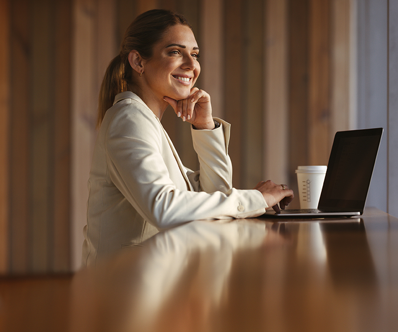 Smiling woman working on a laptop with a coffee cup beside her at a wooden counter.