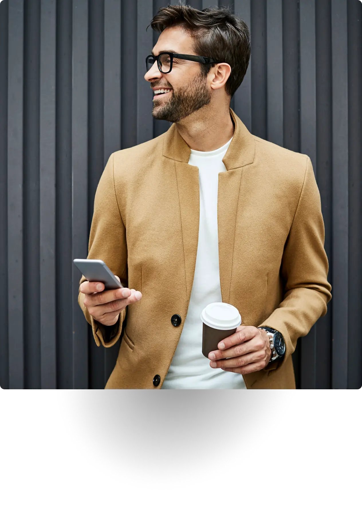 Smiling man in tan coat holding phone and coffee cup.