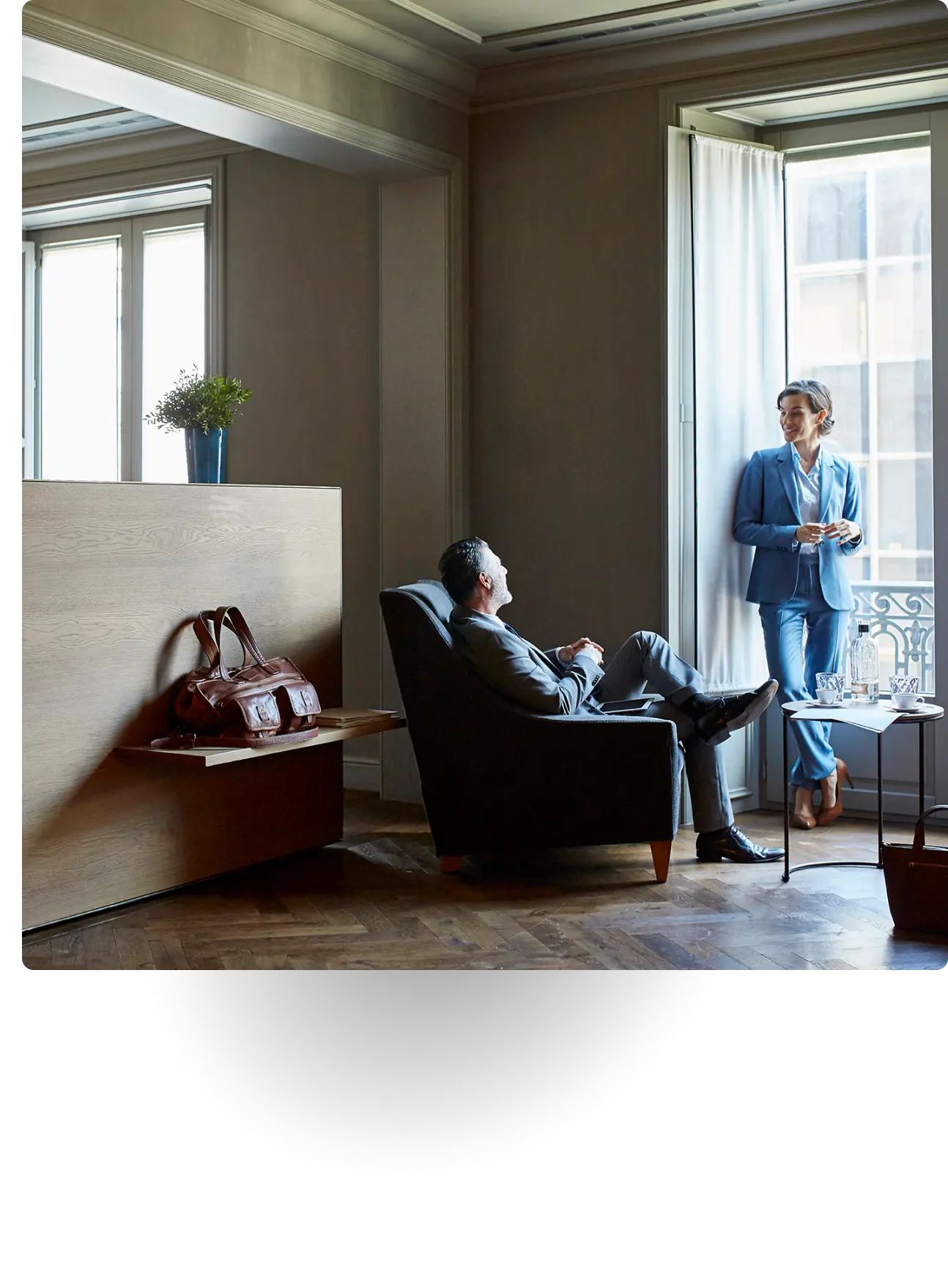 Two business professionals in suits talking near a window in office lounge.