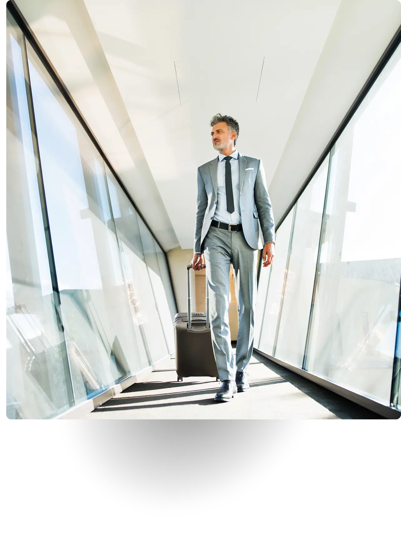 Businessman in suit walking with luggage through glass hallway.