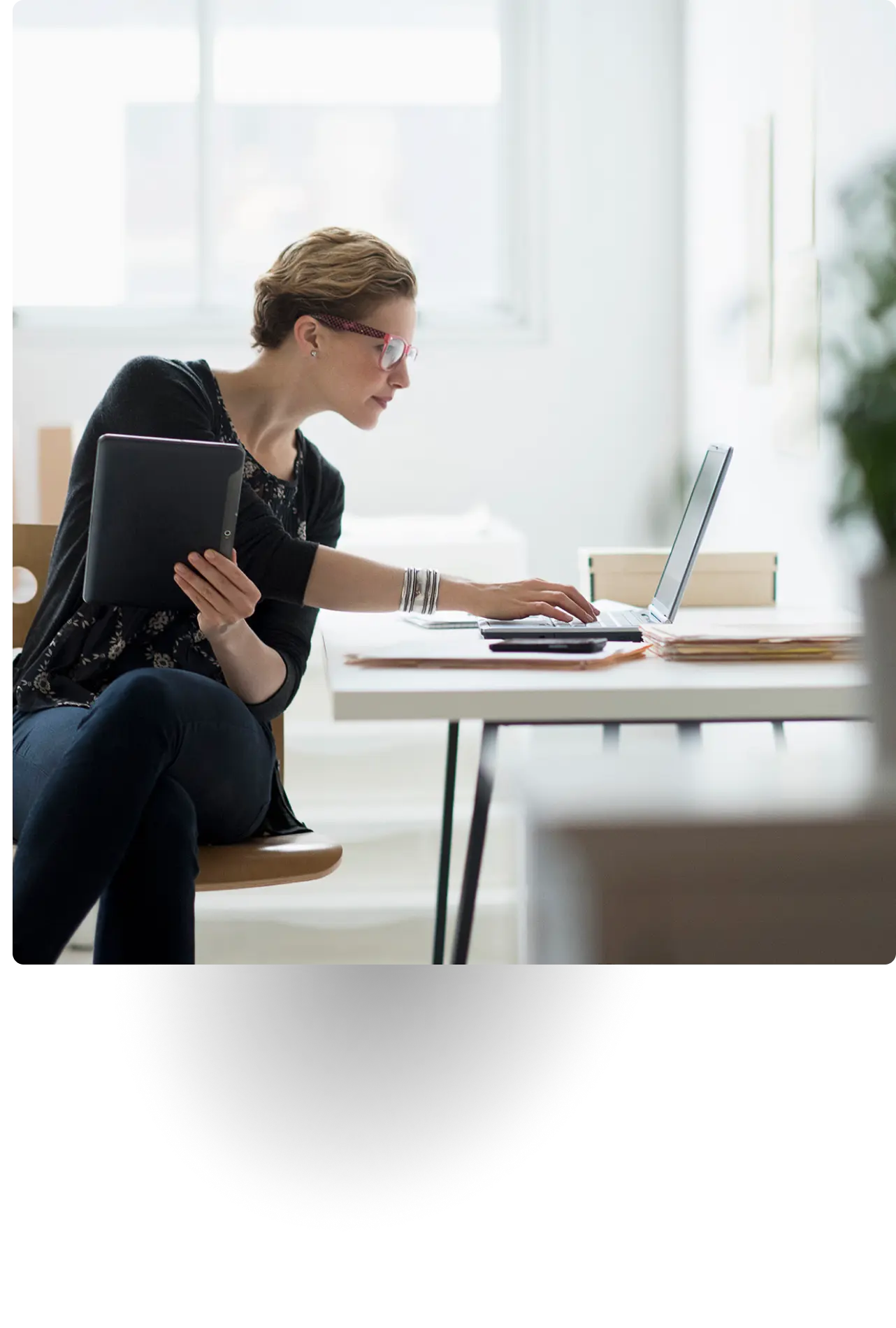 Woman working at a desk with a laptop and holding a tablet in office setting