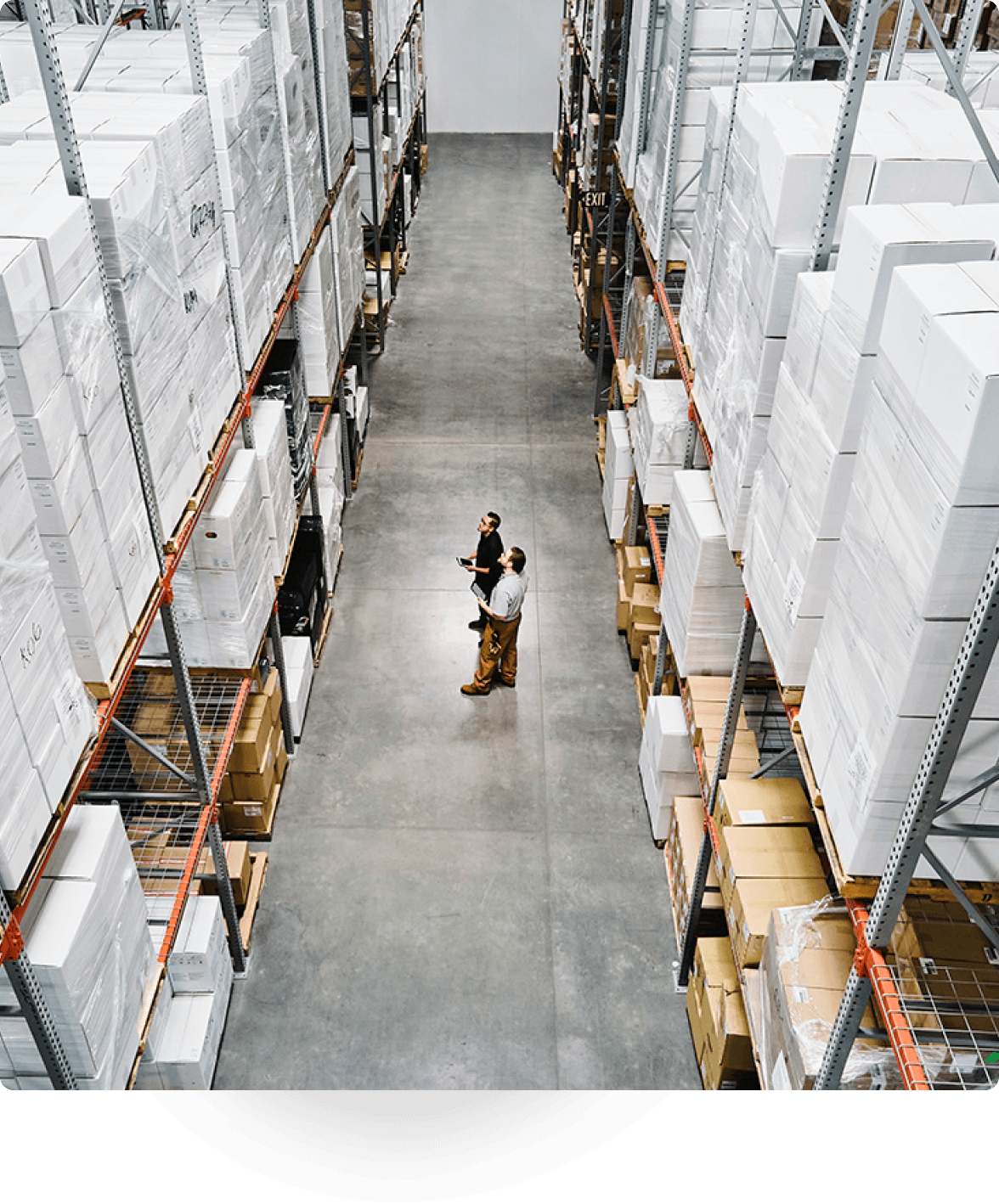 Two people standing in a large warehouse aisle reviewing inventory stacked high on industrial shelves.