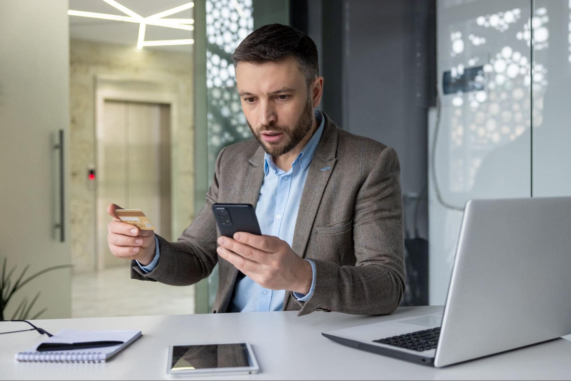 Concerned businessman holding a credit card and checking his phone, representing common signs of financial scams such as unusual payment requests.